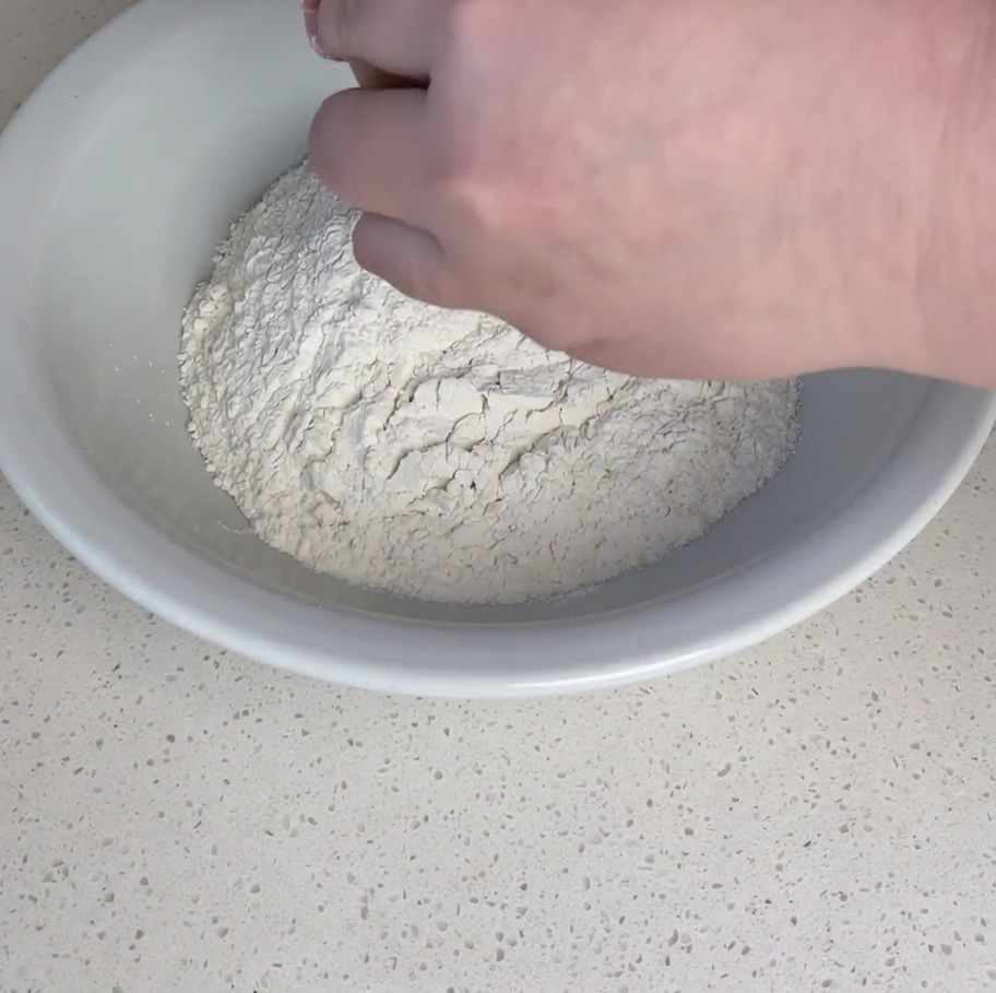 Hands cracking a raw egg into a mound of white high-gluten flour inside a mixing bowl.