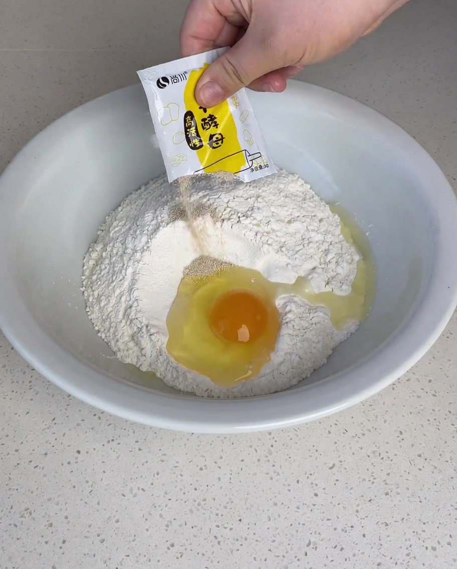 Dry yeast, white sugar, and salt being sprinkled over a mound of flour with a cracked egg in the center.