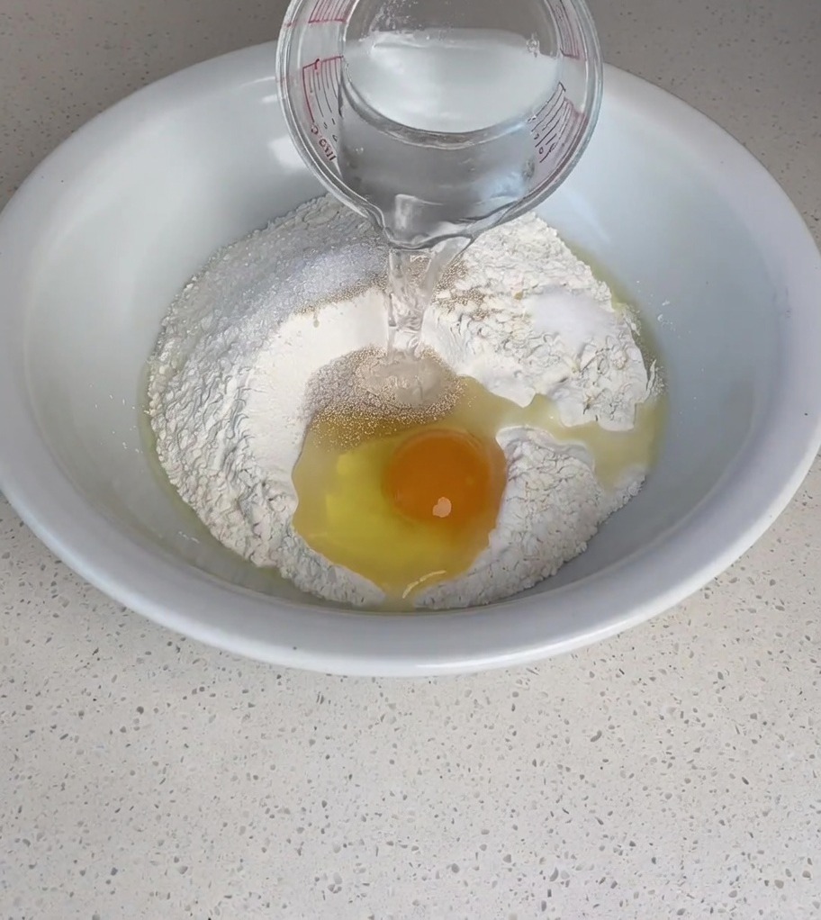 Warm water being poured from a glass measuring cup into a mixing bowl containing flour, an egg, and yeast.