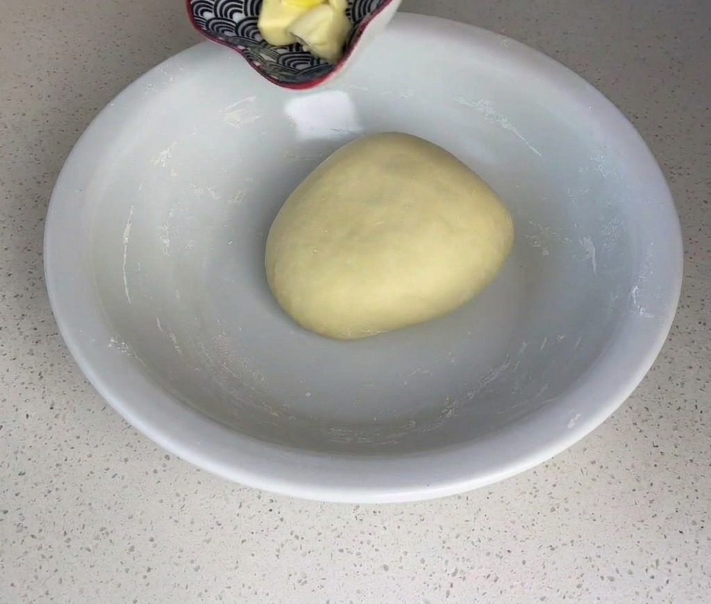 Small cubes of yellow butter being added from a small bowl onto a smooth ball of dough.