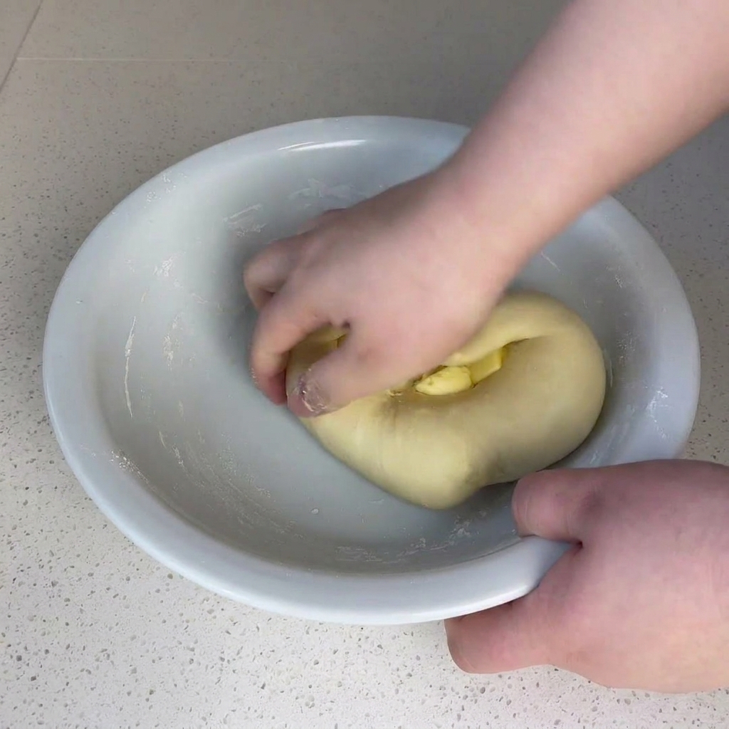 Hands pressing and kneading a small block of butter into a ball of dough inside a large white bowl.