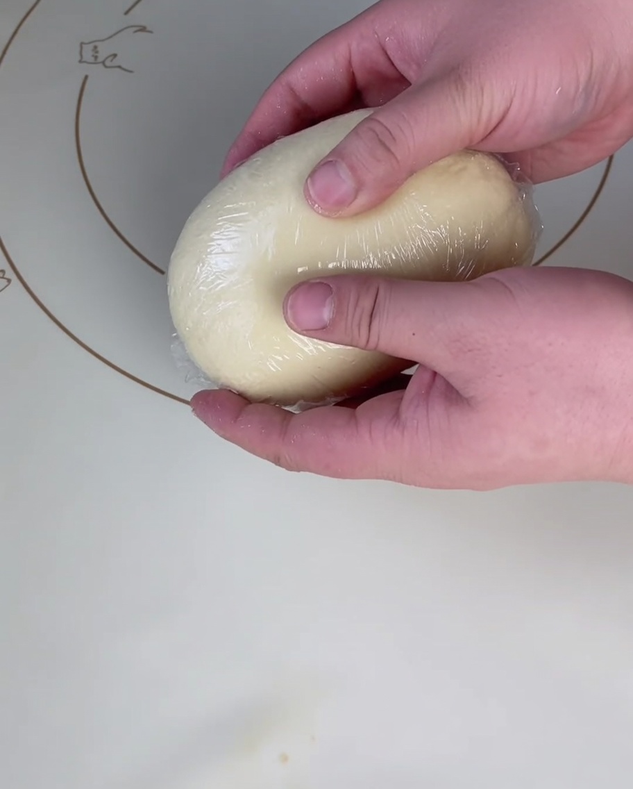 A smooth, round ball of pizza dough being tightly enclosed in a layer of clear plastic wrap on a silicone baking mat.