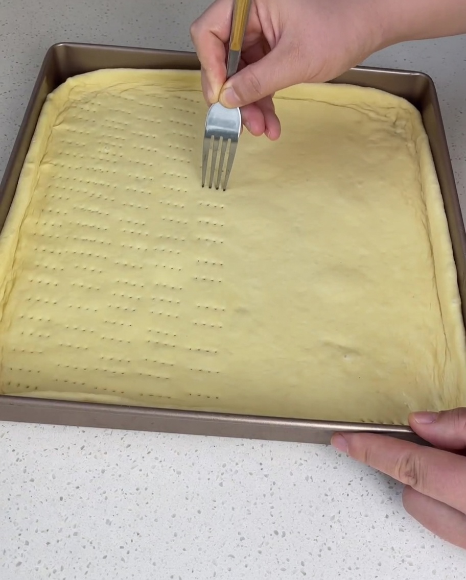 A fork being used to poke multiple rows of small holes into the raw pizza dough inside a square baking sheet.