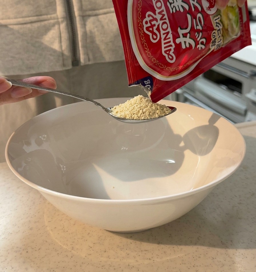 Chicken broth powder being poured from a packet onto a spoon over a large white serving bowl.
