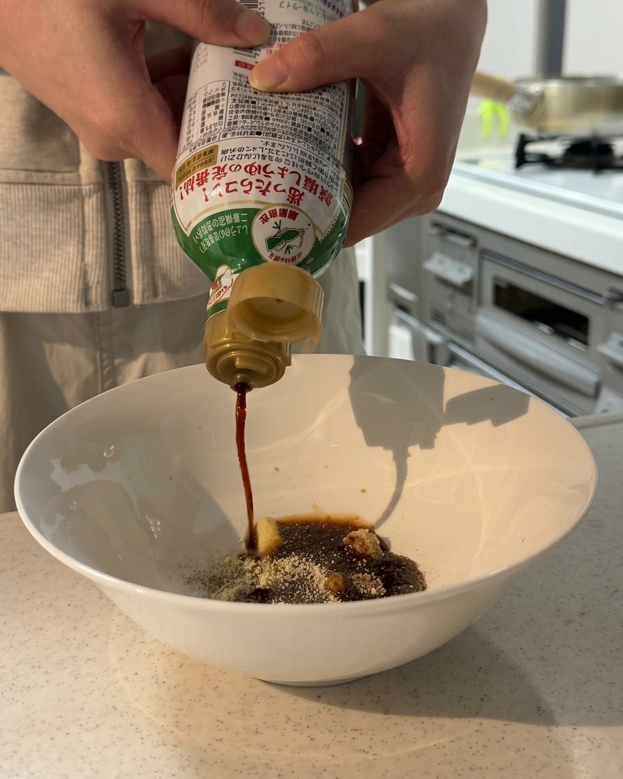 Hands pouring low-sodium soy sauce from a bottle into a white bowl containing dry seasonings and paste.