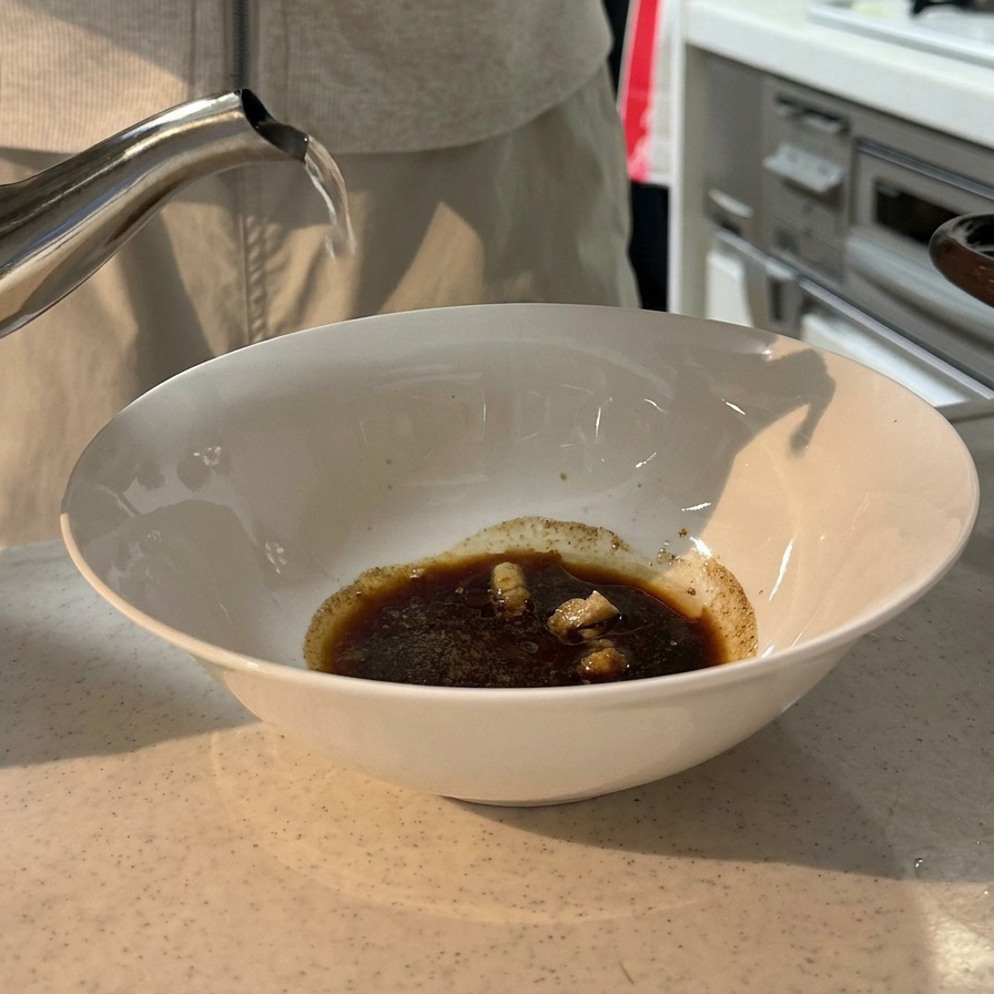Boiling water being poured from a stainless steel kettle into a white bowl containing a dark soy sauce and seasoning mixture.