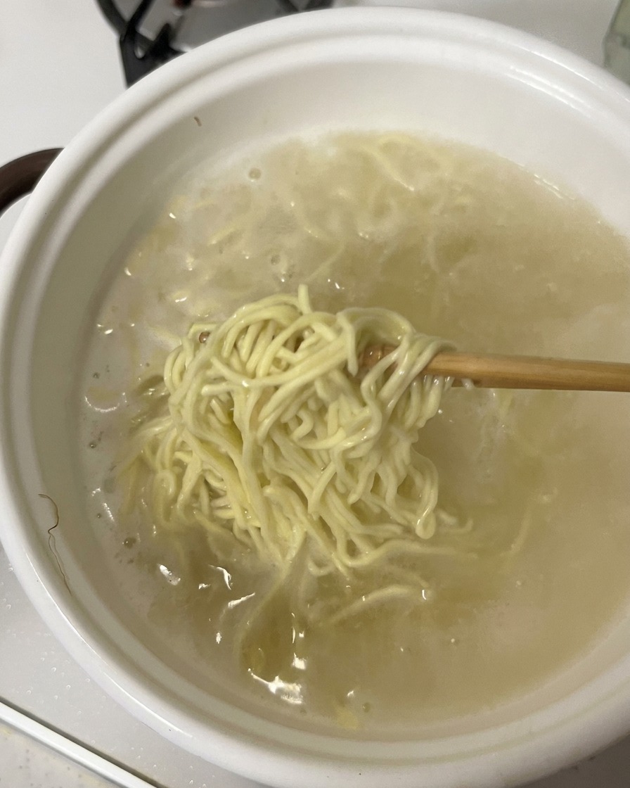 Chopsticks lifting a bunch of cooked ramen noodles out of a white pot filled with boiling water.