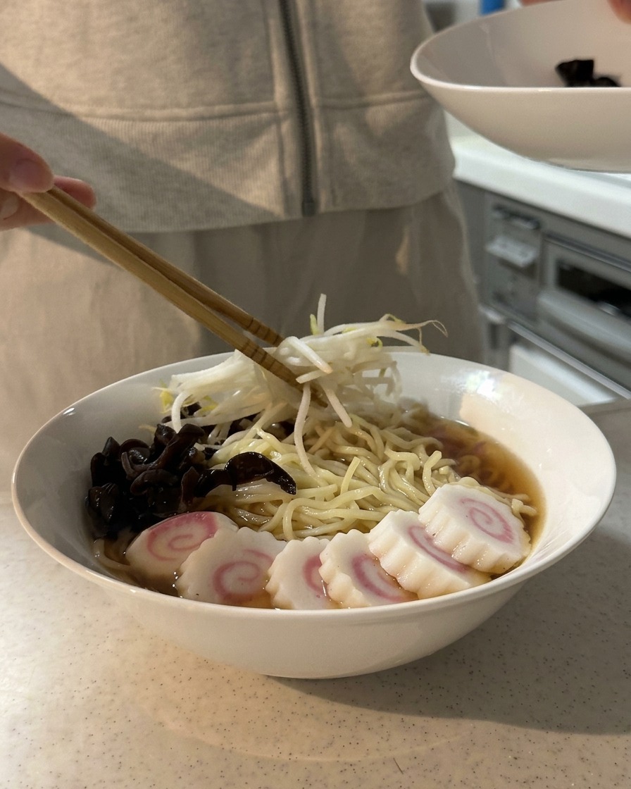 Chopsticks placing a mound of blanched bean sprouts onto a bowl of ramen already topped with wood ear mushrooms and swirl fish cakes.