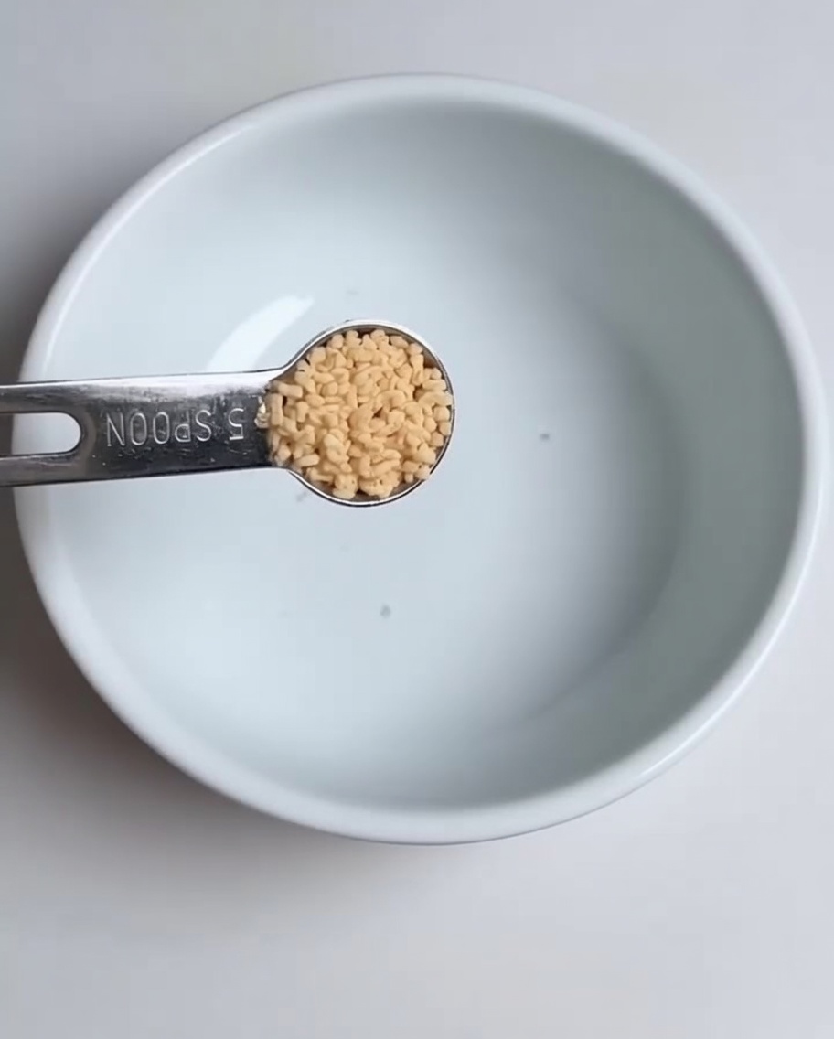 A close-up of a measuring spoon adding chicken bouillon powder into a white bowl.