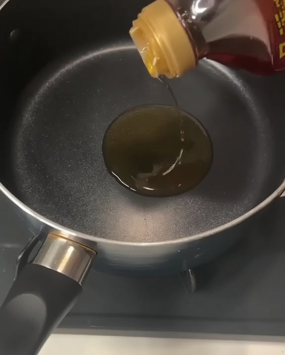 A bottle pouring clear, golden sesame oil into an empty black pot resting on a stovetop.