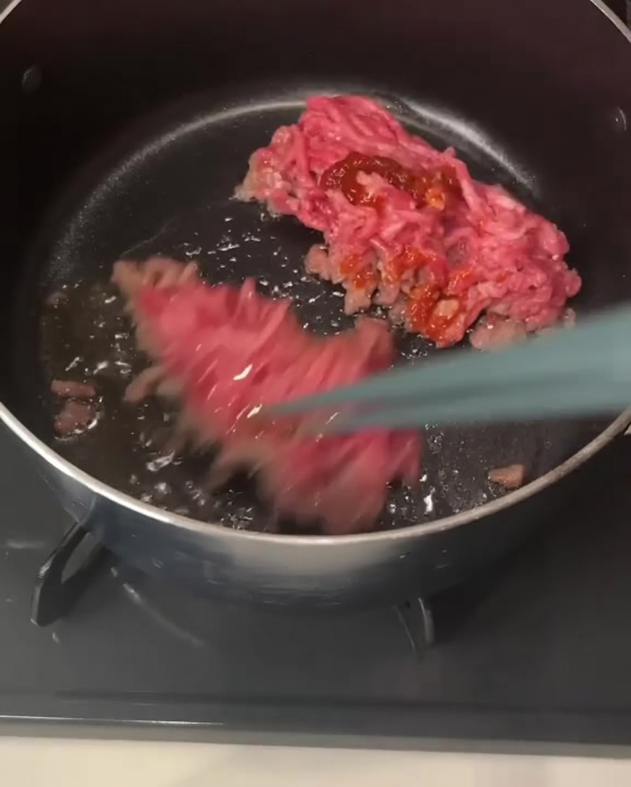 Chopsticks lifting and stirring pieces of ground pork coated in a reddish chili paste inside a pot.
