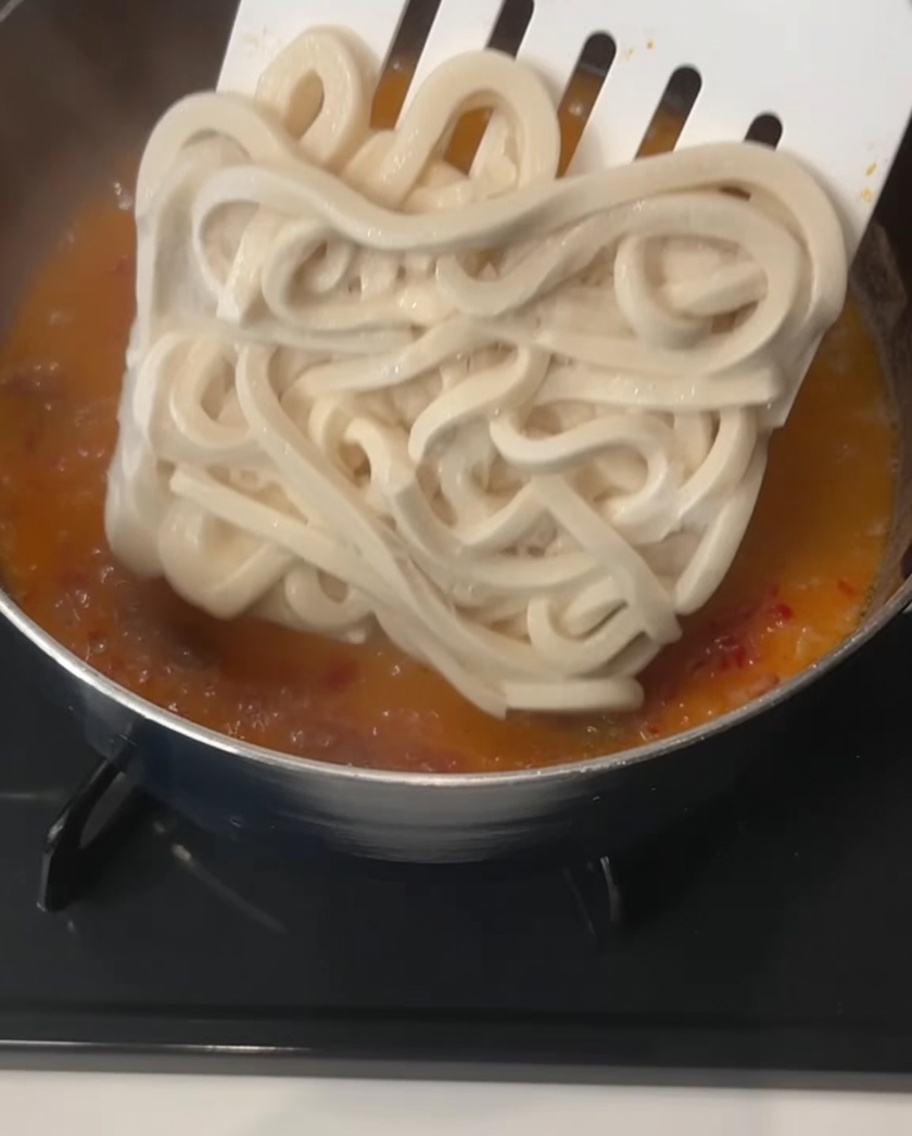 A large, square block of thick frozen udon noodles being lowered by a spatula into a bubbling, creamy orange broth.