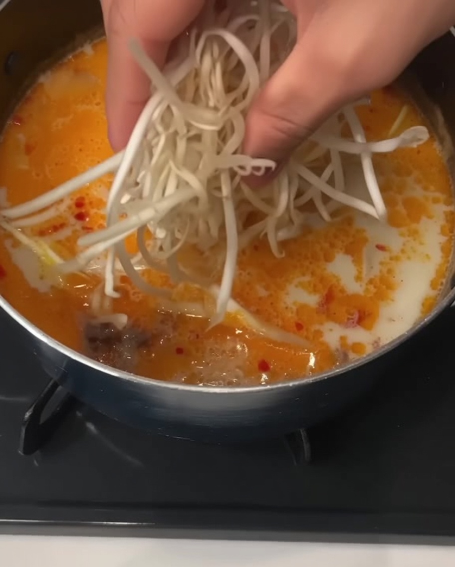 A hand dropping a handful of fresh white bean sprouts into a pot of creamy noodle soup.