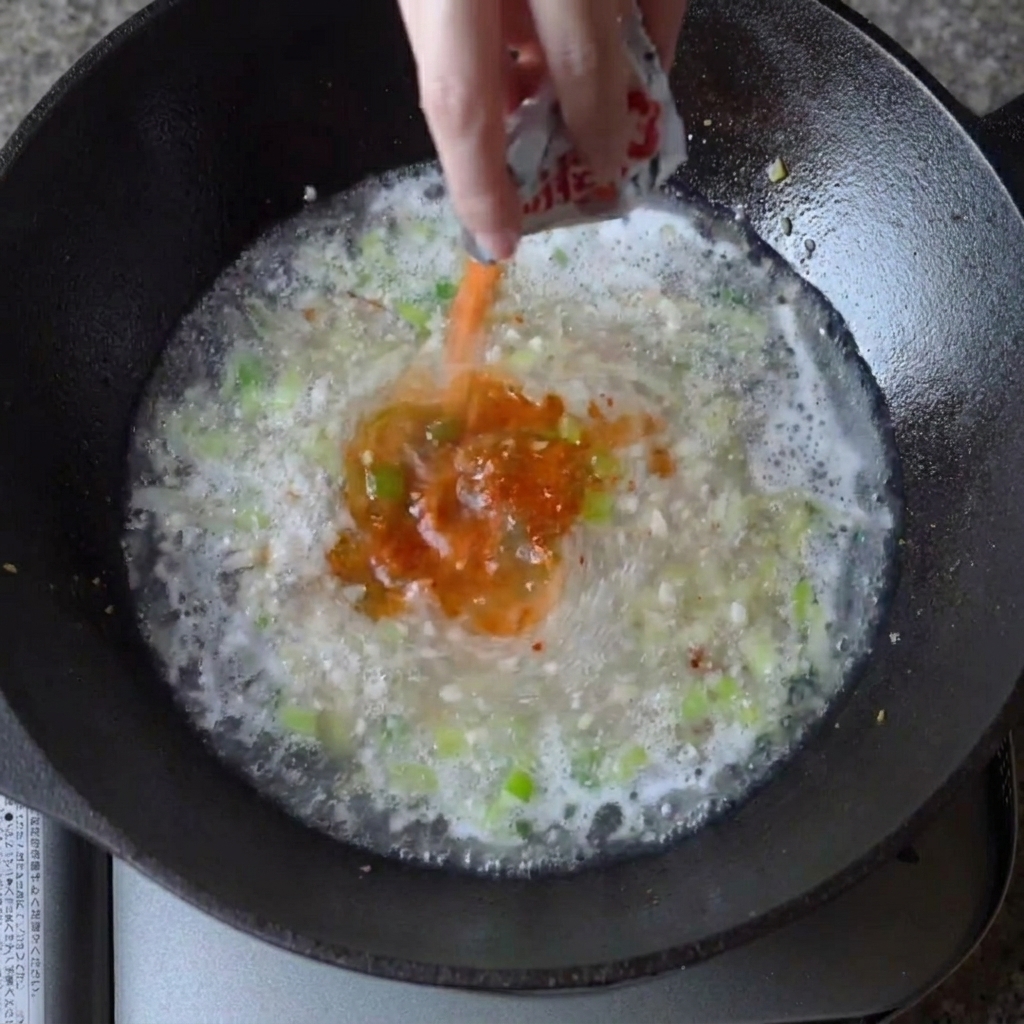 Ramen seasoning powder being sprinkled into the bubbling broth in the wok.