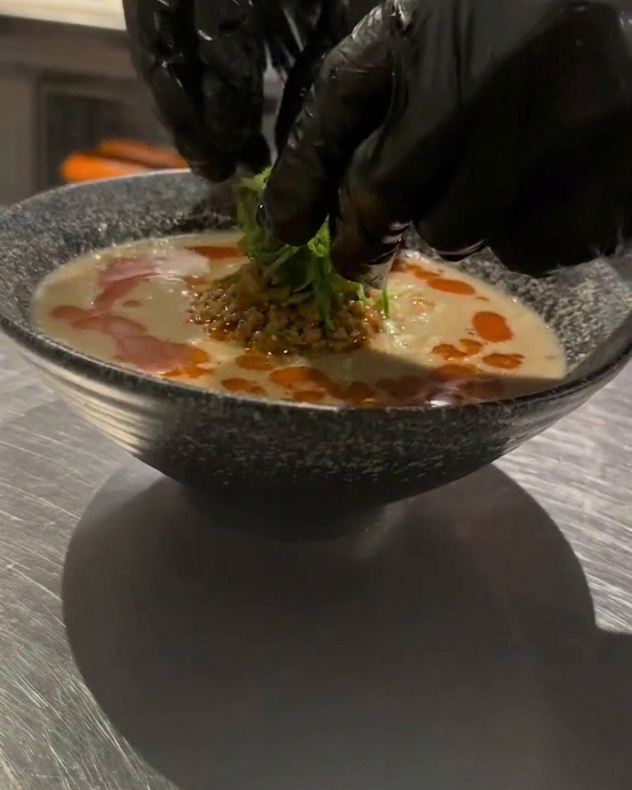 Gloved hands carefully placing shredded green onions on top of a bowl of hot Tan Tan Ramen as a final garnish.