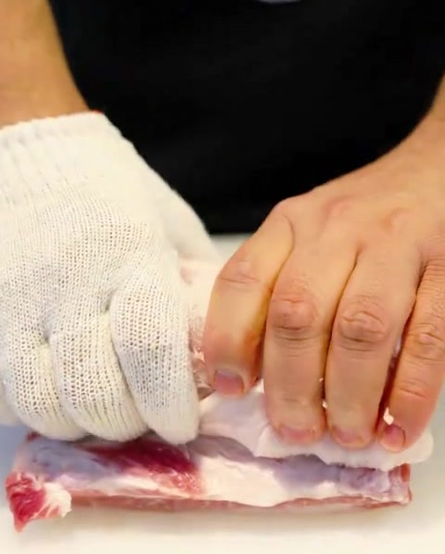 A raw pork belly roll being tightly bound with kitchen twine by hands wearing protective gloves.