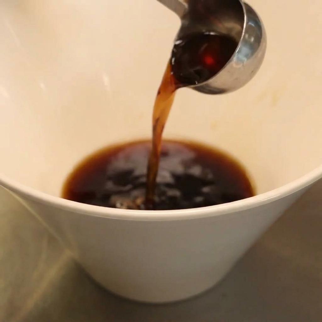 Dark liquid seasoning being poured from a metal ladle into a deep white bowl.
