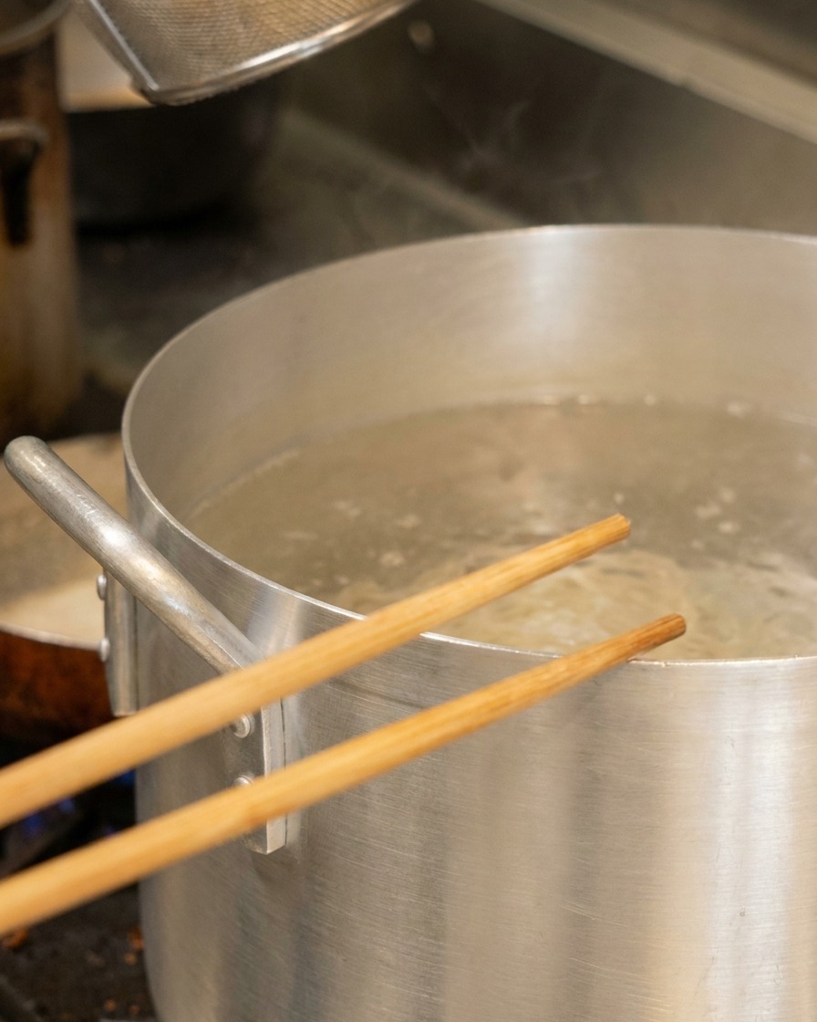 Wooden chopsticks resting over a large aluminum pot of boiling water containing a noodle basket.