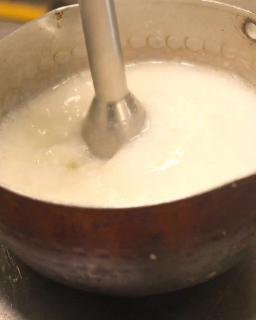 A hand blender being used to create a creamy foam in a pot of chicken paitan soup.