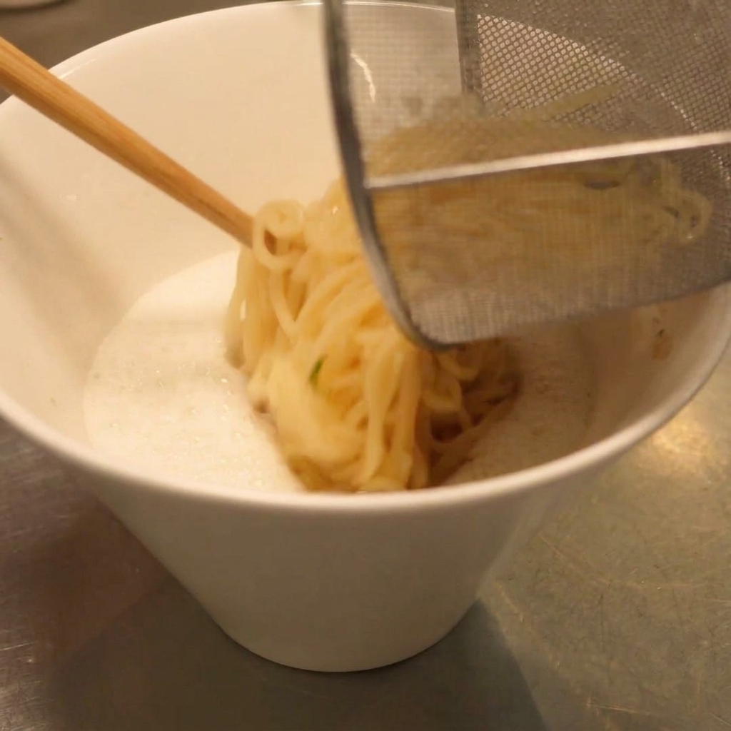 Drained ramen noodles being transferred into the bowl of creamy soup using a kitchen basket.