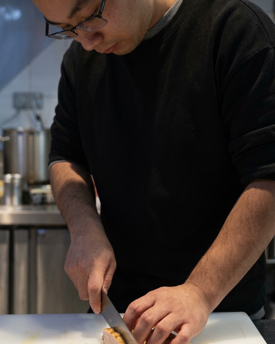A sharp knife cutting through cooked chicken chashu on a white cutting board.