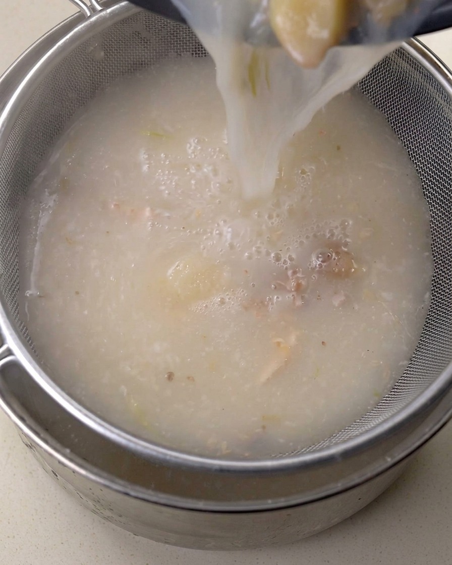 Rich broth being poured through a fine metal strainer.