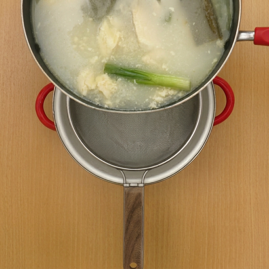 Hot chicken broth being poured through a metal sieve into a clean pot.