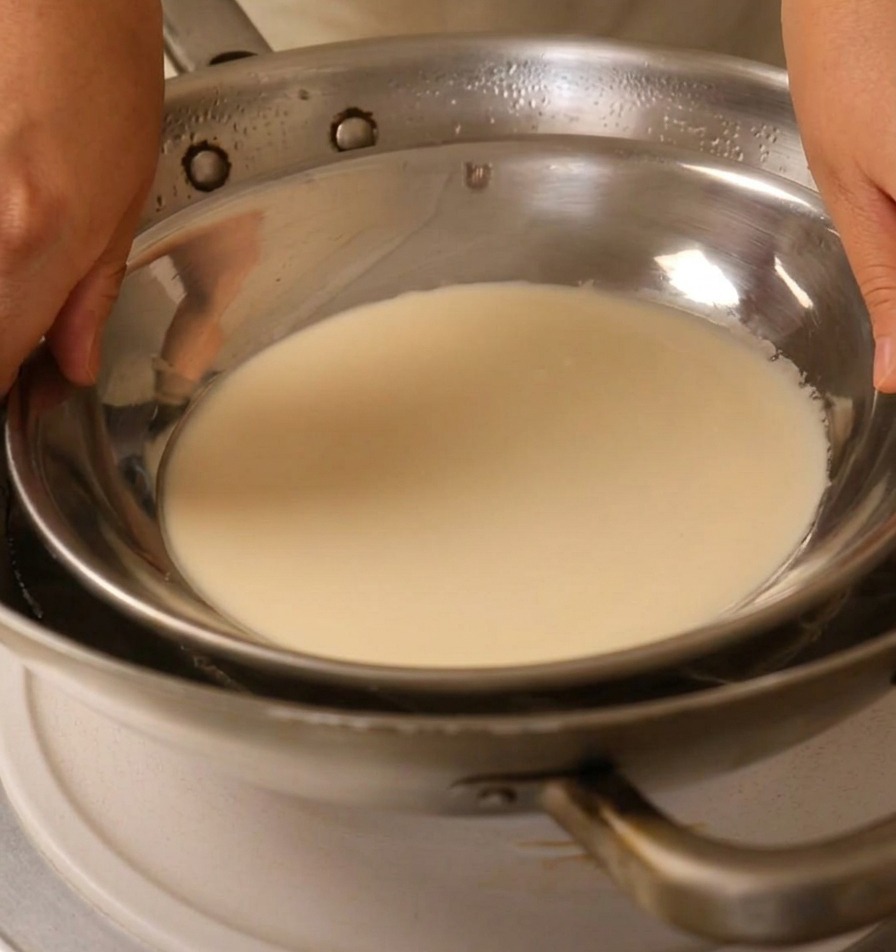 Hands placing a metal plate filled with white batter over a wok of boiling water.