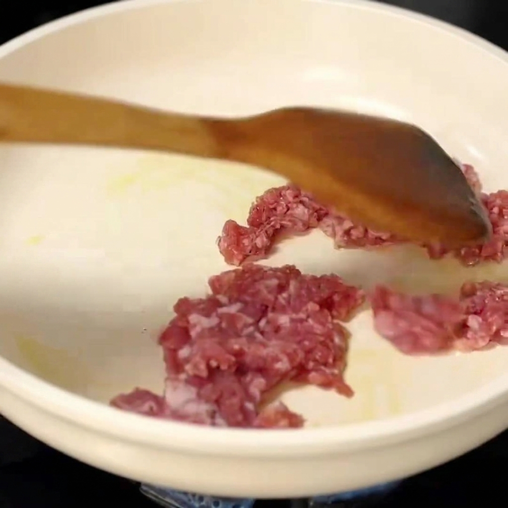 Raw minced pork being broken apart and stir-fried in a white pan with a wooden spatula.