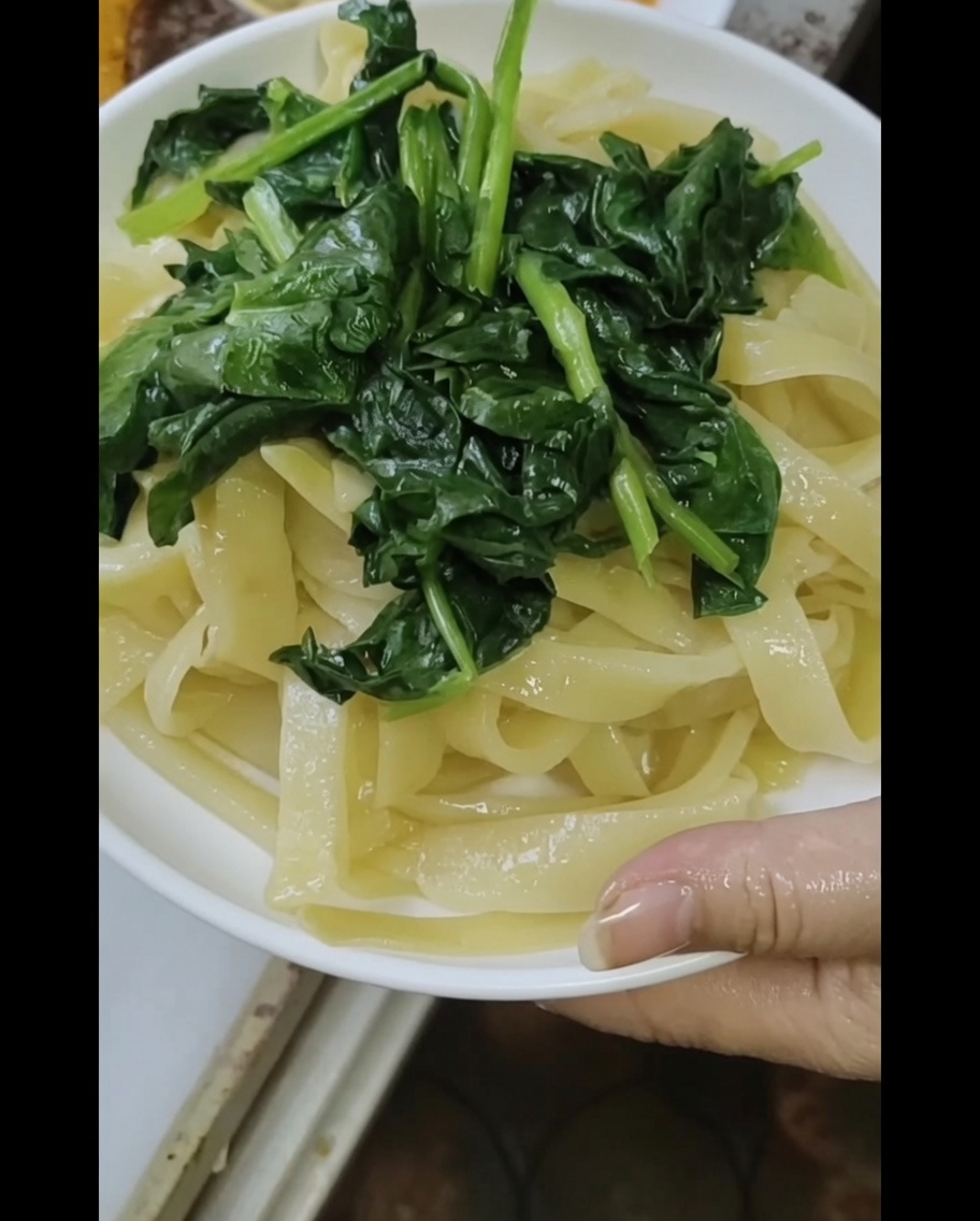 A close-up view of freshly steamed noodles topped with blanched spinach, being drizzled with a rich sesame and peanut sauce.