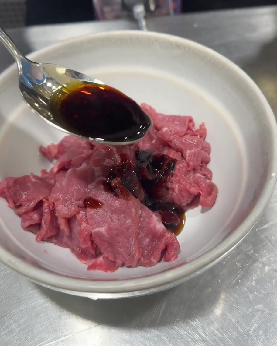 Sliced raw beef in a bowl being seasoned with soy sauce, salt, pepper, and cornstarch.