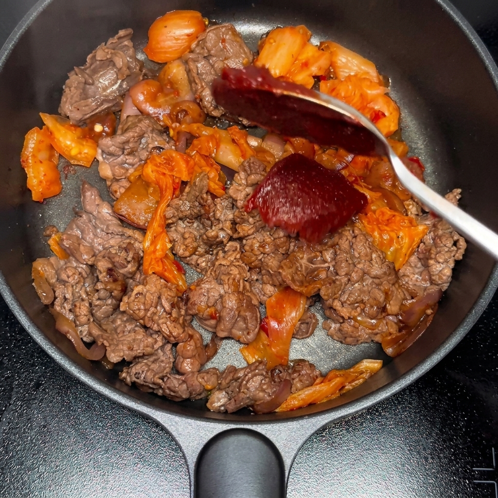 Korean chili paste and chili powder being added to the stir-fried beef and kimchi mixture.