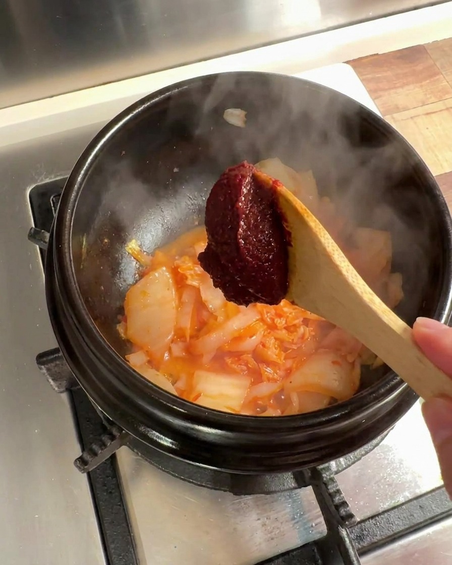 A spoonful of Korean chili paste being added to the mixture of kimchi and onions in the stone pot.