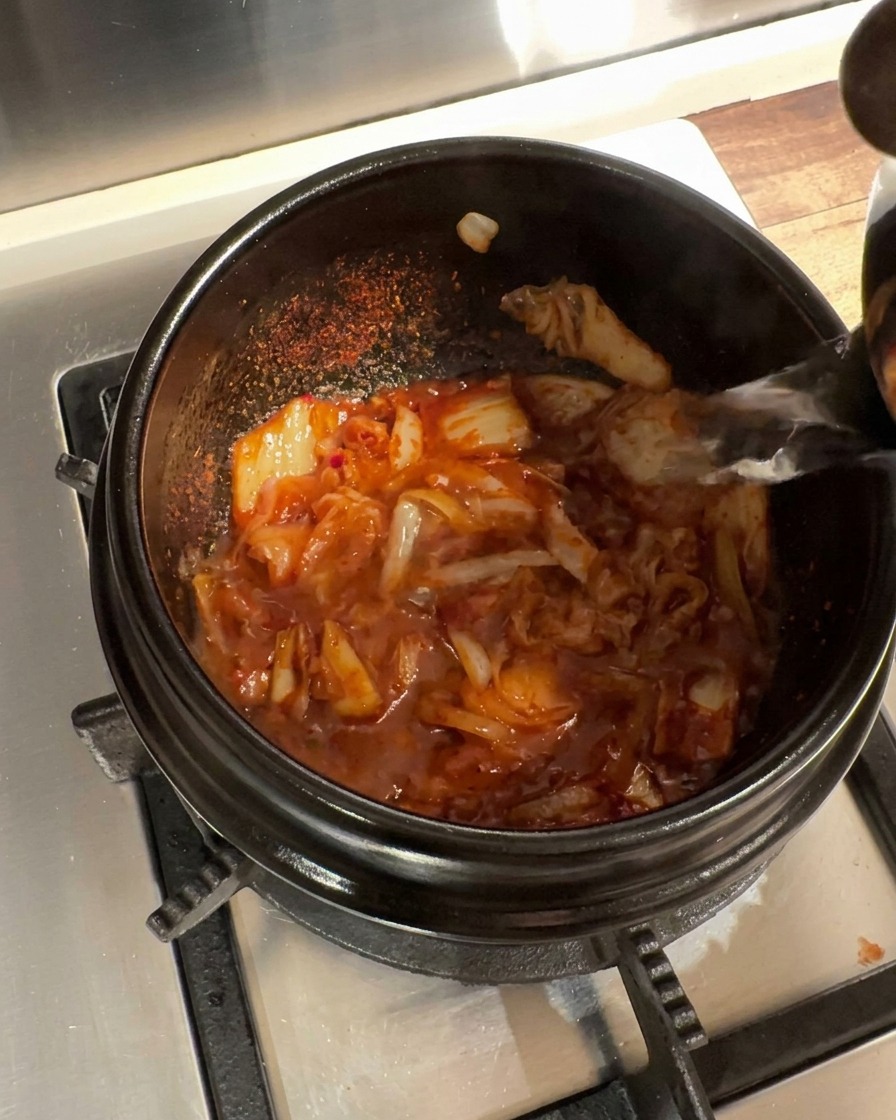 Hot water being poured into the stone pot containing the seasoned kimchi and onion mixture.