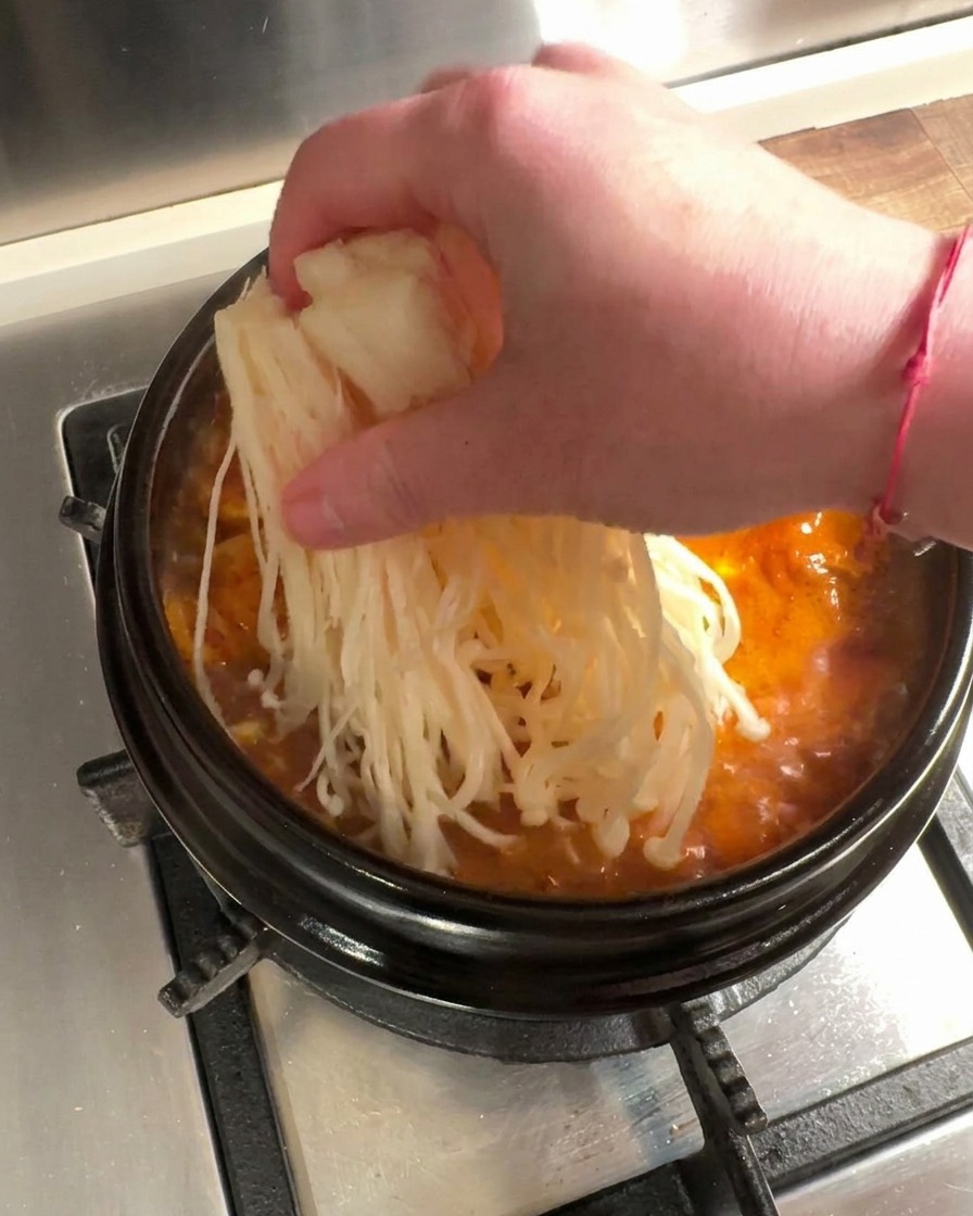Fresh enoki mushrooms and thinly sliced raw beef being placed into a bubbling red kimchi stew.