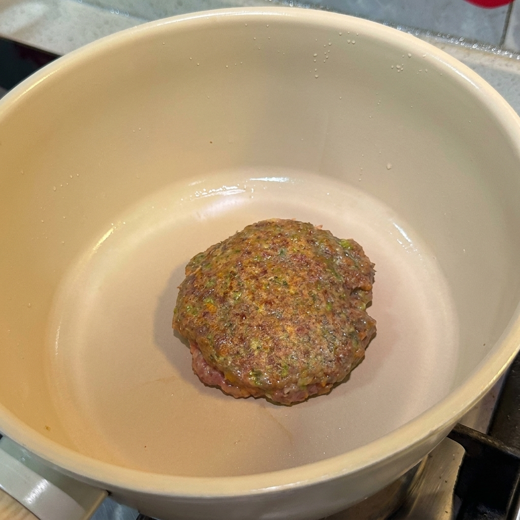 A seasoned beef patty cooking in a pan, showing a browned surface.