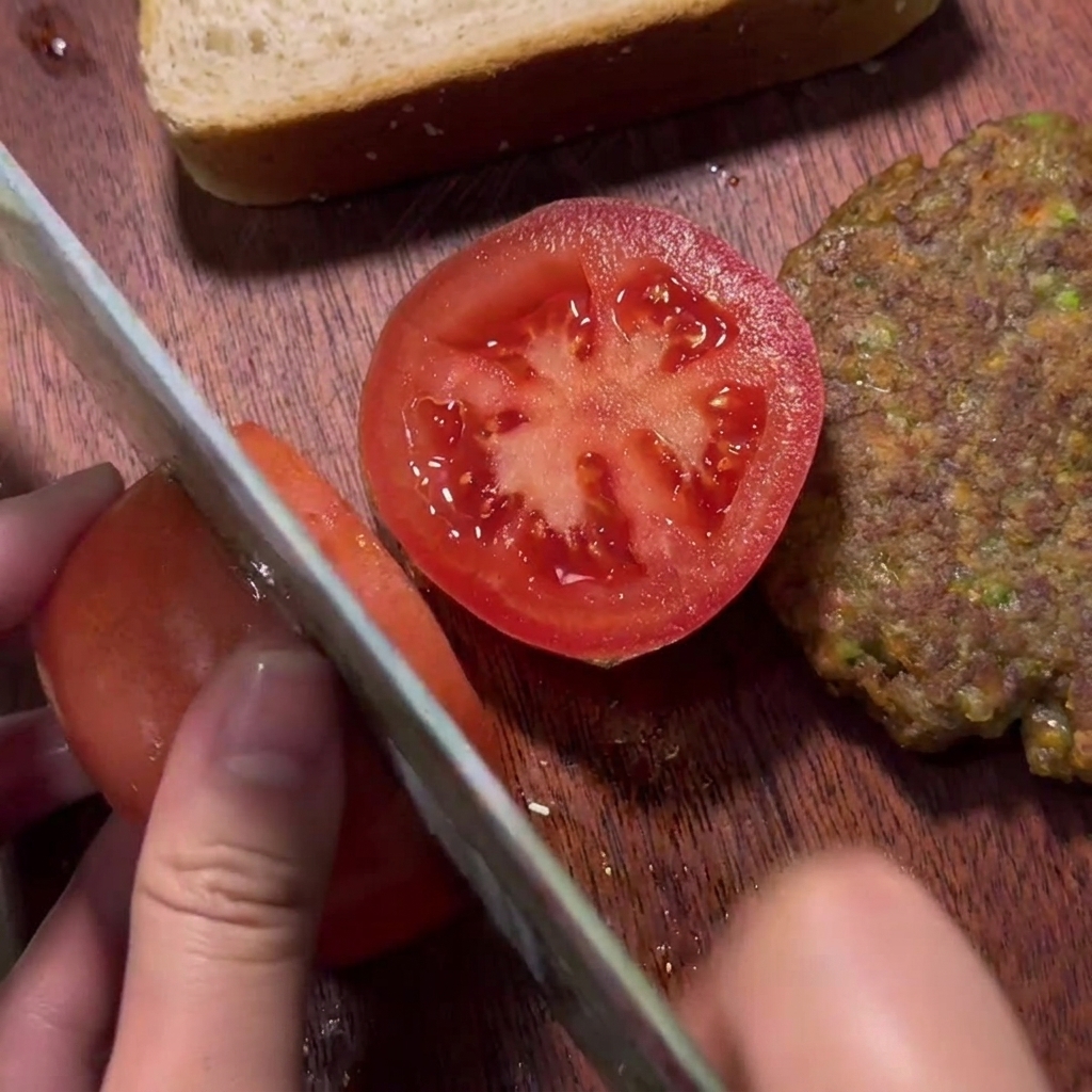 A hand slicing a fresh red tomato on a wooden board next to a cooked beef patty.