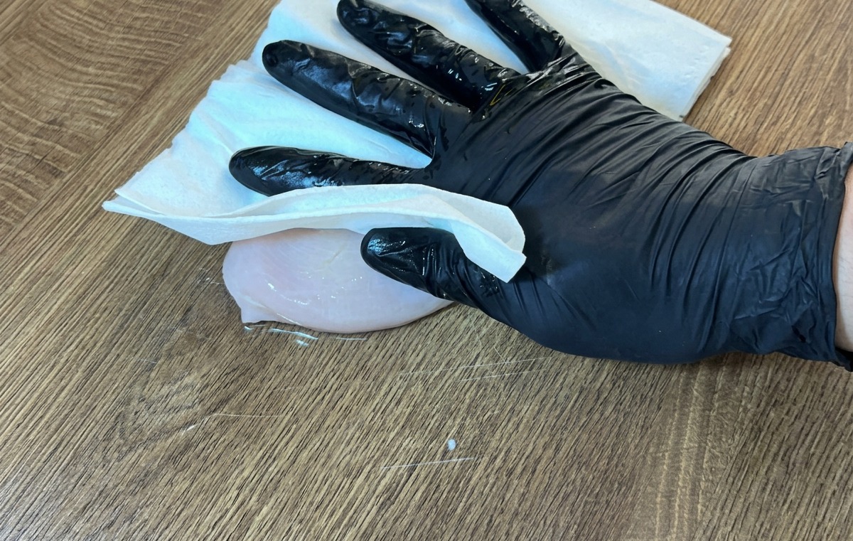 A gloved hand using a paper towel to pat a raw chicken breast dry on a wooden surface.