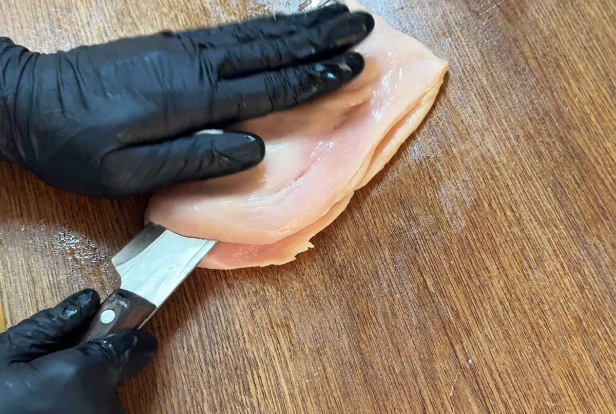 A sharp knife slicing horizontally into a chicken breast on a cutting board to butterfly it.