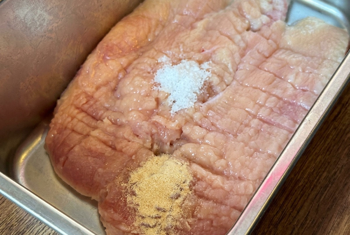 A raw chicken breast in a metal tray covered with various dry seasoning powders and sauces.