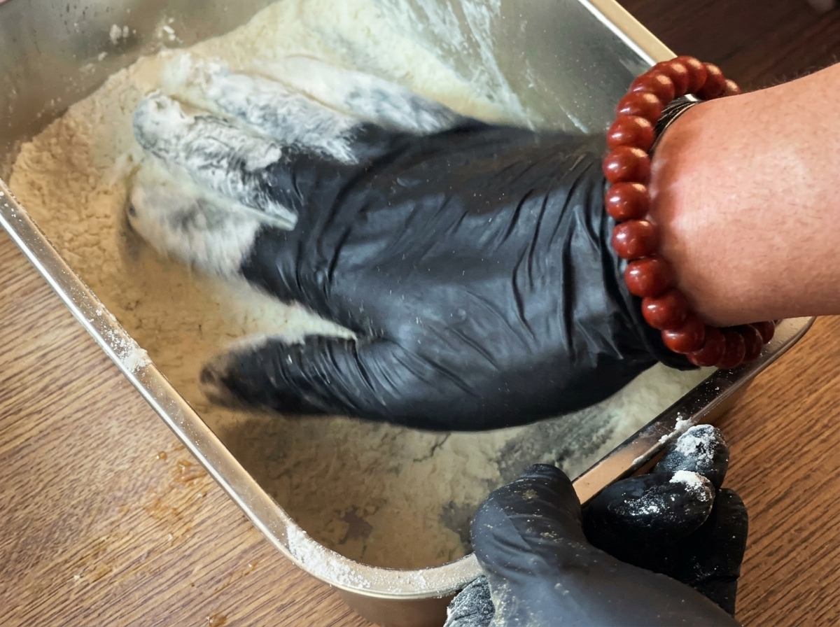 Stuffed chicken breast being coated in white starch in a metal tray.