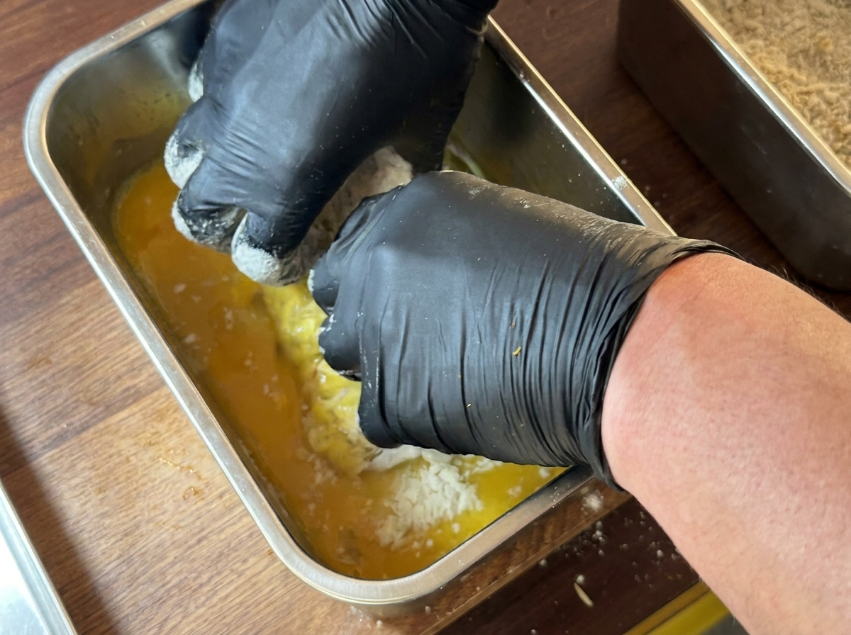 A chicken breast piece being fully submerged in a container of yellow egg wash.