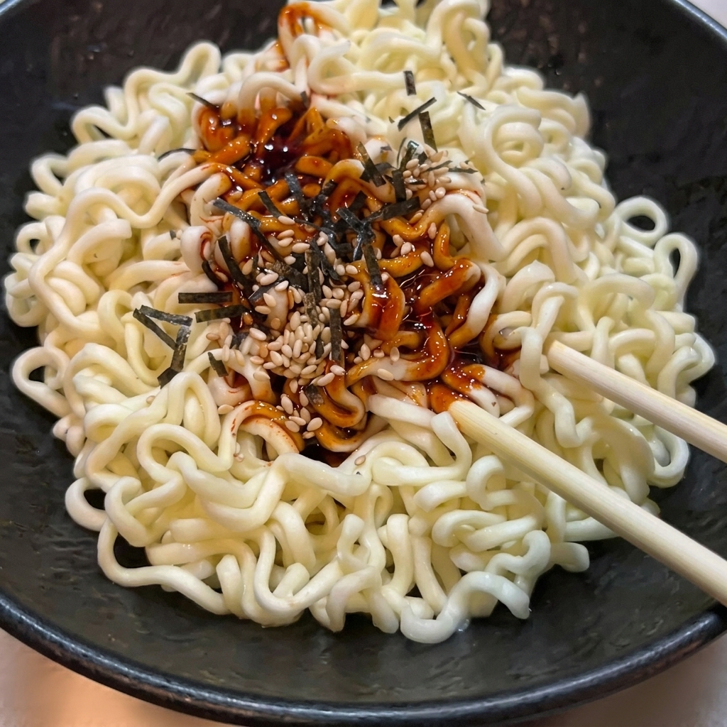 Cooked wavy ramen noodles in a textured black bowl being tossed with bright red spicy sauce, sesame seeds, and seaweed using wooden chopsticks.