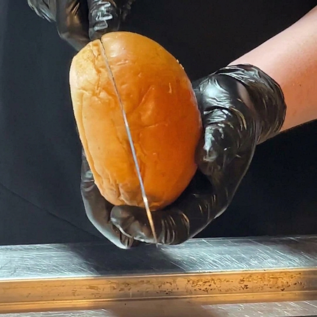 Hands wearing black cooking gloves using a long knife to slice a shiny brioche burger bun in half.