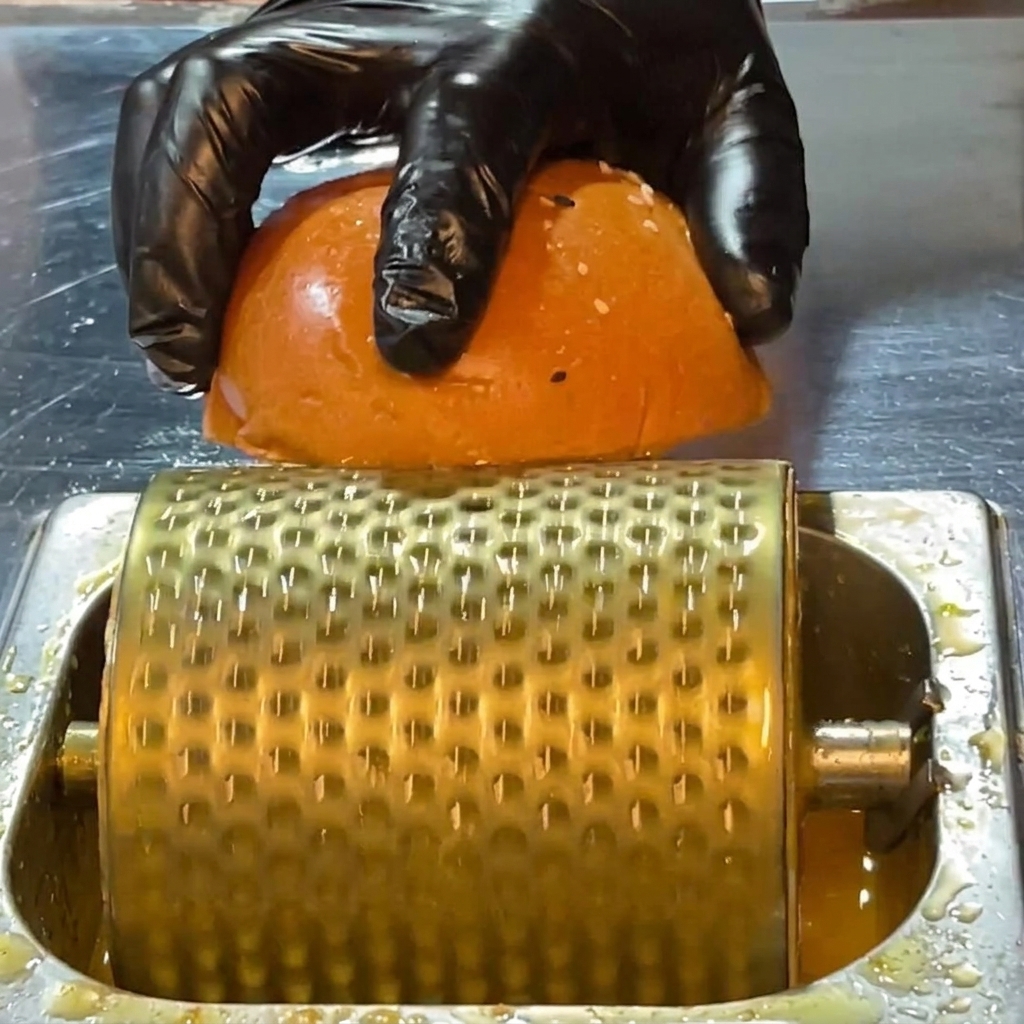 A gloved hand pressing a burger bun half over a perforated metal butter roller sitting in melted butter to coat the cut side.