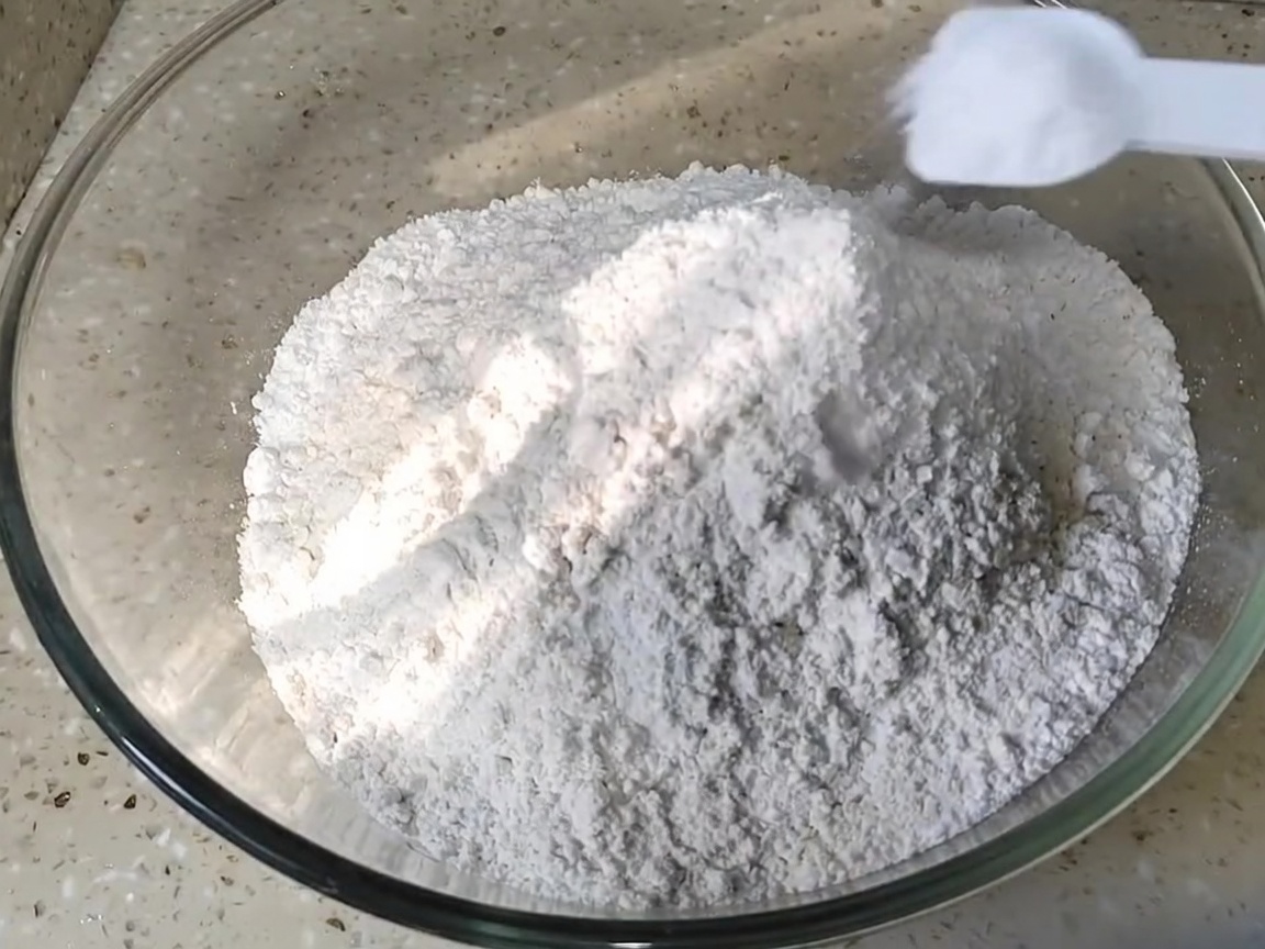 A spoonful of salt being added to a mound of white flour in a glass mixing bowl.