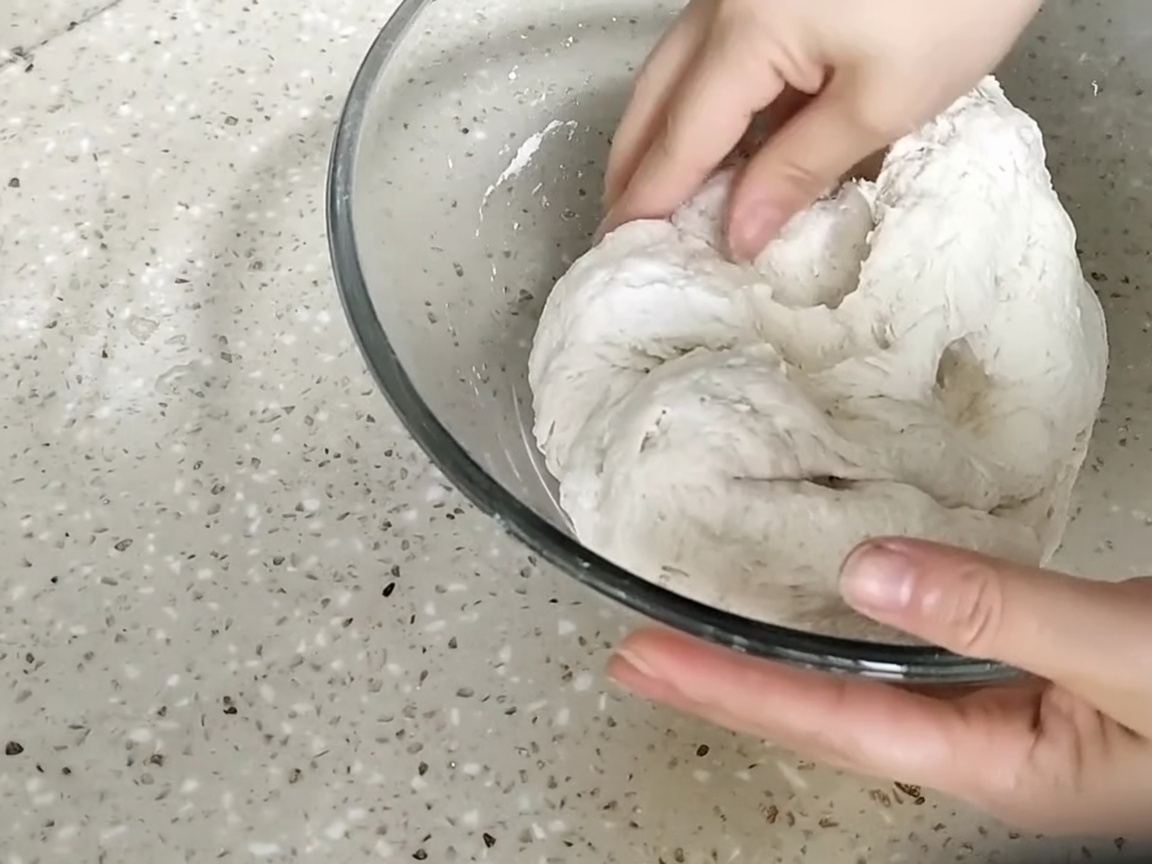 Hands pressing and kneading a rough, shaggy ball of dough inside a glass bowl.
