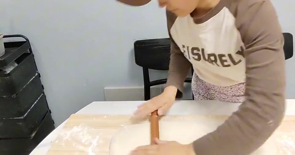 A person using a wooden rolling pin to flatten a block of dough on a floured wooden board.