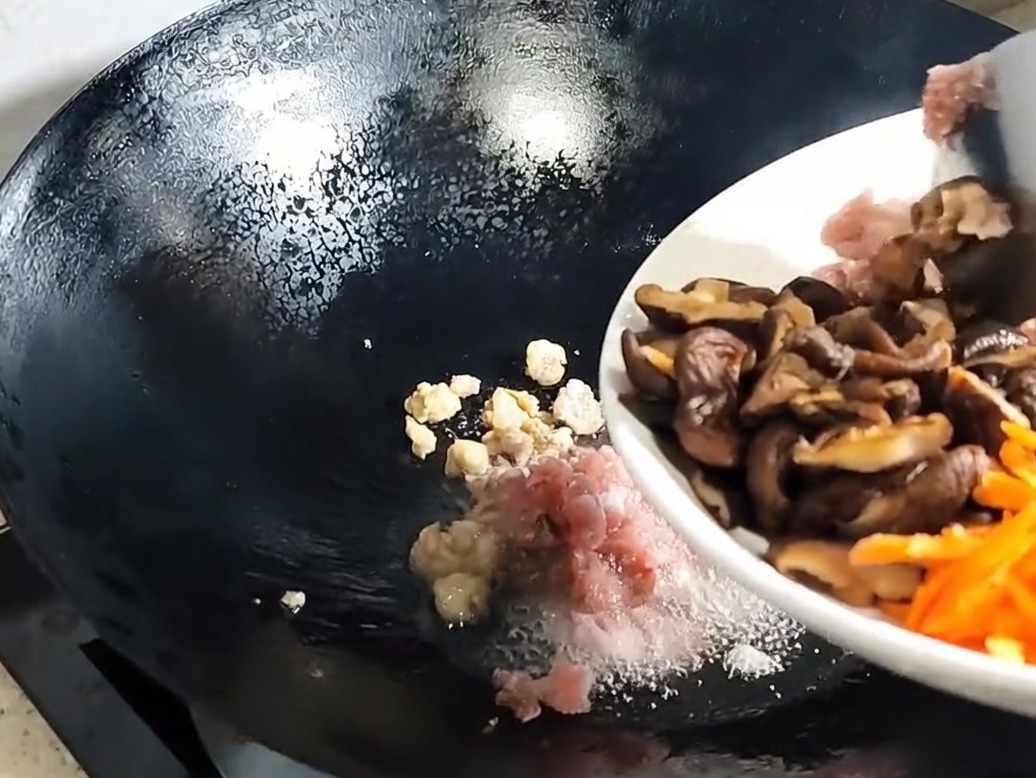 Sliced shiitake mushrooms and aromatics being added to the rendered pork in the wok.