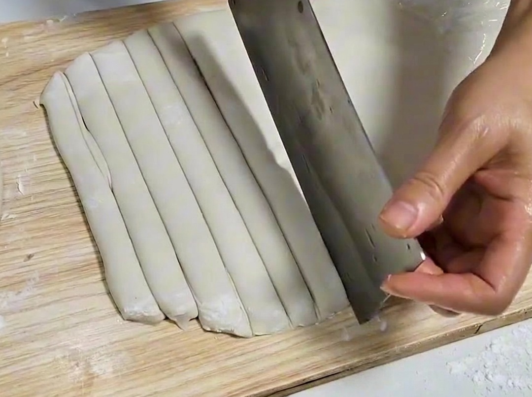 Dough sheet being sliced into even strips with a metal cleaver on a wooden board.